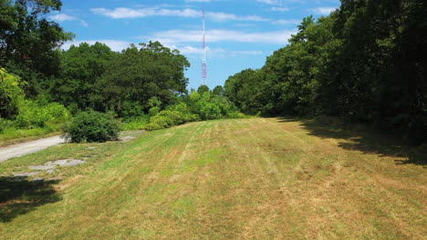 pull-away-down-a-tree-lined-path,-away-from-a-tall-red---white-antenna-in-the-distance,-on-a-sunny-day-with-beautiful-blue-skies-with-light-white-clouds