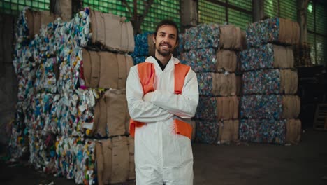 Portrait-of-a-confident-and-happy-brunette-man-with-a-beard-in-a-white-protective-suit-and-an-orange-vest-who-stands-near-recycled-plastic-at-a-waste-recycling-plant