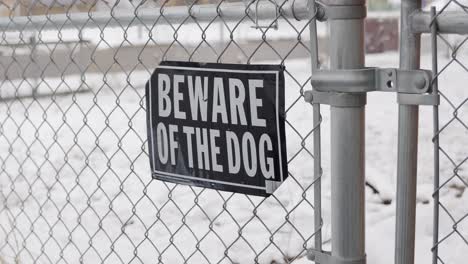 Snowing-on-first-day-of-spring-as-camera-zooms-in-and-out-of-Beware-Of-The-Dog-sign-on-chain-link-fence-with-black-lab-dane-labradane-dog-in-yard-with-lots-of-snow-on-ground---in-Cinema-4k-60-fps