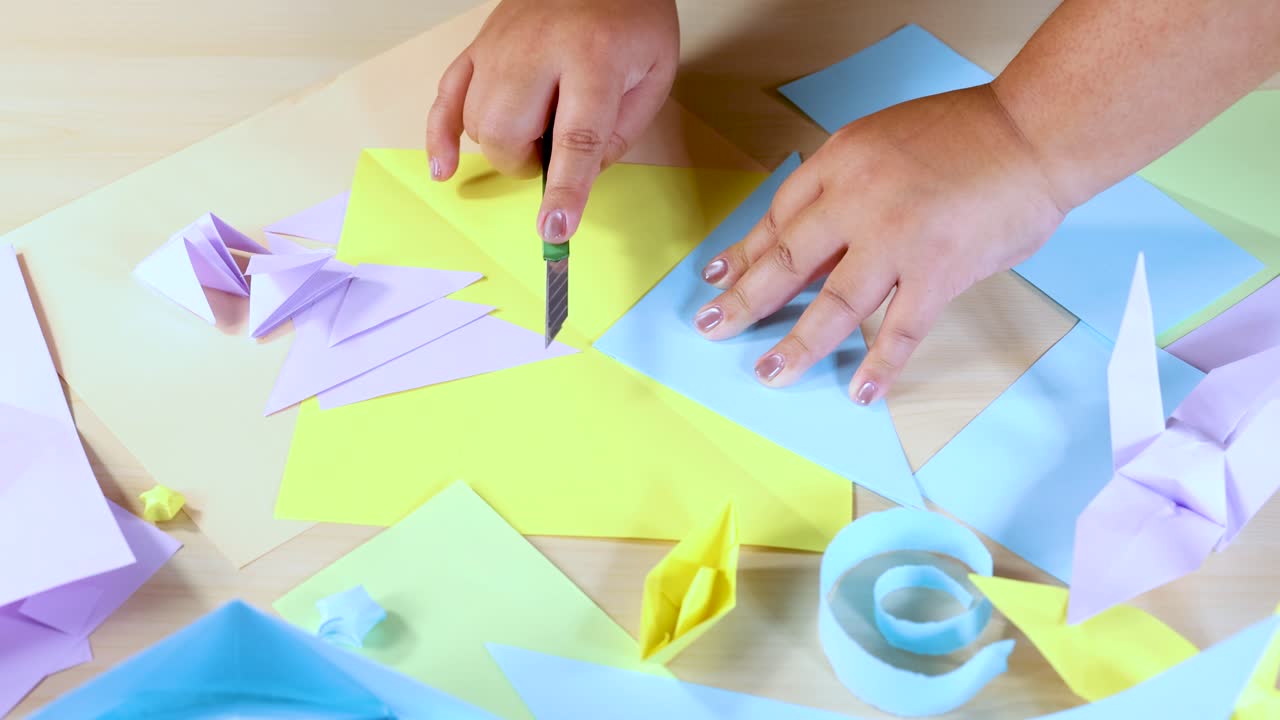 Person cuts and folds pastel origami paper on wooden table under bright, even lighting