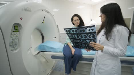 woman doctor radiologist explains the results of ct scanning for young female patient, showing the snapshot with images, observing and analyzing ct scan in modern clinic beside modern ct scanner