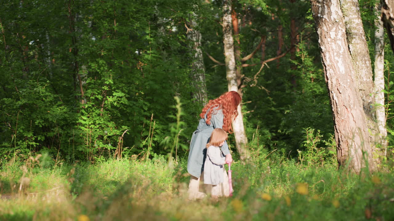 Long shot of little girl riding scooter with help of mother through green forest path under soft sunlight, both enjoying playful outdoor moment filled with warmth, laughter, and connection in nature