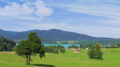 Wonderful-view-at-the-turquoise-colors-of-the-popular-Tegernsee-in-the-southern-bavaria---zoomed-out-with-the-great-view-over-a-green-meadow-under-a-blue-sky-with-mountains-in-the-background