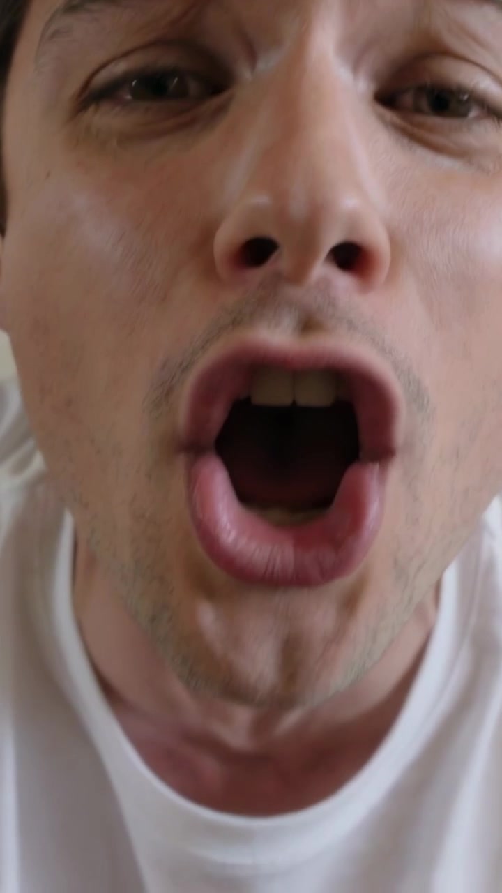 Vertical video: Leaning man making faces, shouting on beige couch, camera starting close, white tee