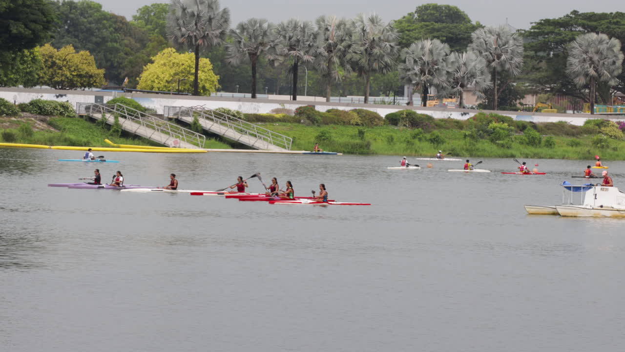 People kayaking on calm water with trees and buildings in the background, training for competition during overcast day