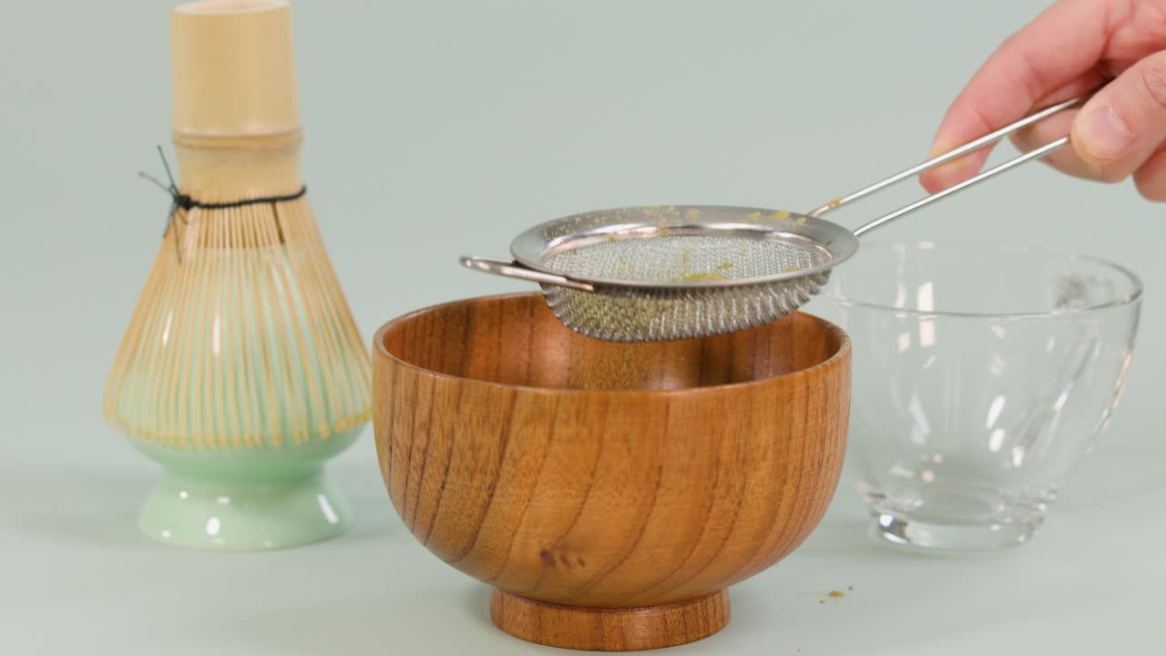 Hand sifts matcha powder through strainer into wooden bowl, with bamboo whisk and glass cup