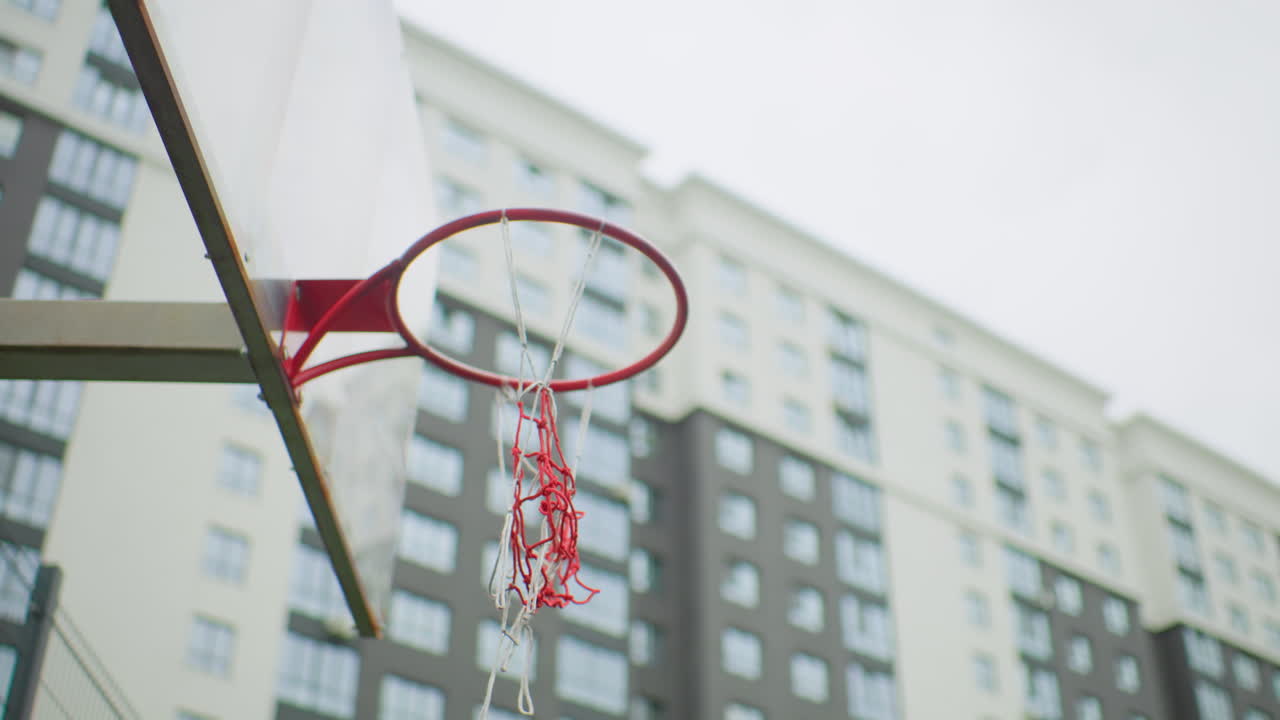 Canestro da basket urbano con pallone sospeso sopra il cerchio, rete a catena strappata che oscilla sullo sfondo di un edificio residenziale sotto un cielo nuvoloso, campo in cemento vuoto, tonalità tenui, effetto mosso che cattura un tiro sbagliato