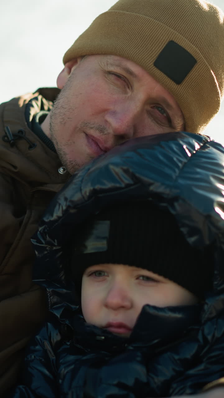 отец и сын сидят на скамейке, с головой отца, опирающейся на голову сына, с отцом, держащим сына близко, оба смотрят сфокусировано, с размытым видом деревьев на заднем плане