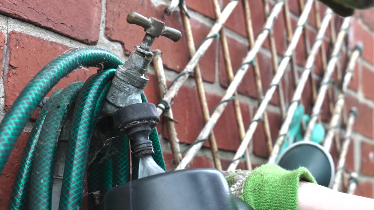 Ręka w rękawiczce odkręca kran ogrodowy, gdy woda wlewa się do plastikowej konewki obok zwiniętego węża na ceglanej ścianie, ukazując prace w ogrodzie, nawadnianie i rutynę domowego ogrodnictwa