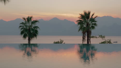 Escena-Del-Resort-Al-Atardecer-Piscina-Con-Palmeras-Mar-Y-Montañas