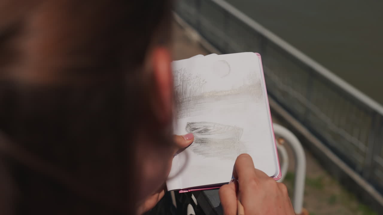 sketchbook drawing on riverside railing outdoors, artist hands shading pencil over graphite study of river and bridge, sunlit evening mood, closeup view, gentle breeze, urban leisure moment