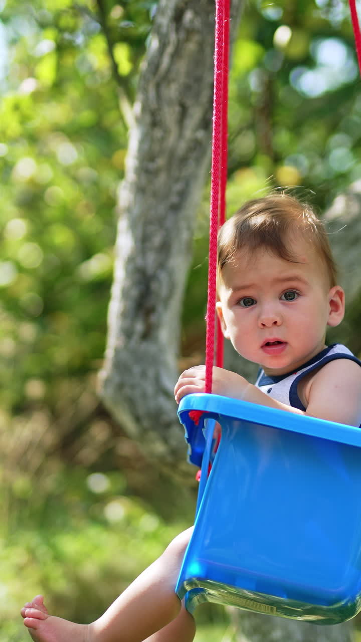 Lovely toddler swinging on a blue swing in the garden. Baby looking attentively into camera. Side view. Blurred backdrop. Vertical video