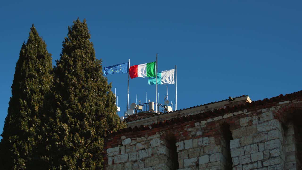 Bandiere d'Europa, Italia e della regione Lombardia che sventolano contro un cielo azzurro limpido al Castello di Brescia, slow motion