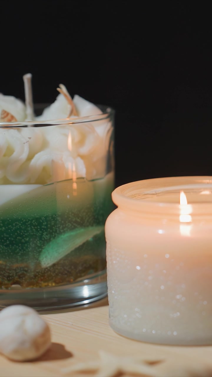 Nærbillede af tændt lys med blødt flakkende flamme i glasbeholder ledsaget af dekorative elementer som muslingeskaller og lagdelte lys. Voksens tekstur er synlig under varm belysning