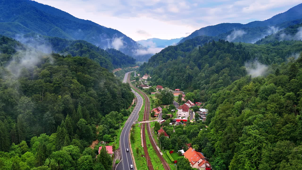 Mountain valley with road and village surrounded by forest, Romania. Aerial panorama of a road, railway, and small village nestled between green forested mountains in the Carpathians, Romania