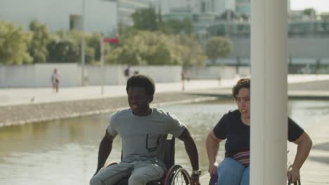 Happy-multiethnic-friends-using-wheelchairs-walking-at-river