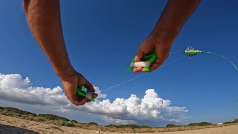 Vista-En-Primera-Persona-De-Las-Manos-Masculinas-Que-Controlan-La-Cometa-Verde-Voladora-Con-Cola-Larga-Sobre-La-Playa-De-Arena-Sosteniendo-Manijas-Verdes