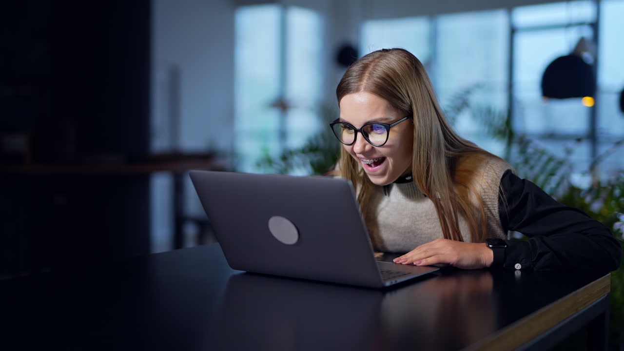 Female employee opens laptop and exclaims in surprise. Happy young woman cheering, smiling and gesturing in excitement. Blurred background.