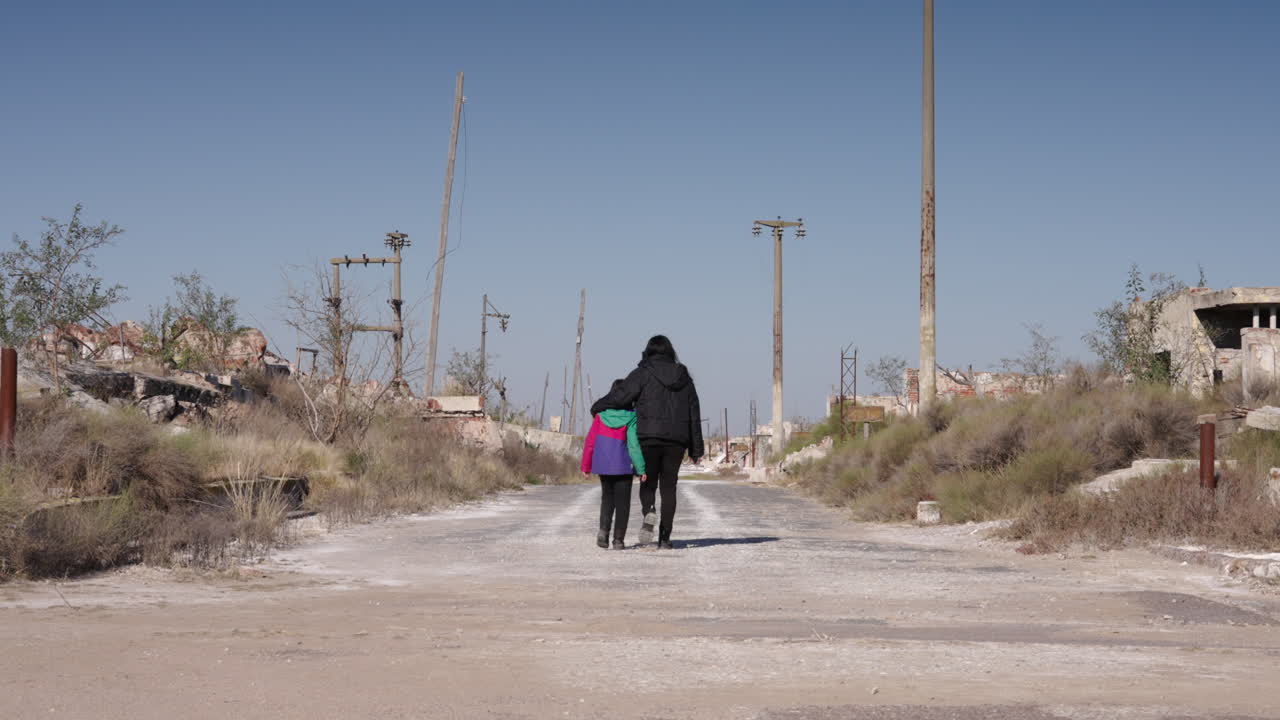 A mother and daughter walk away through the ruins of Epecuén, Argentina. Light poles line the weathered road, creating vanishing point under a sunny sky with faded, cinematic colors