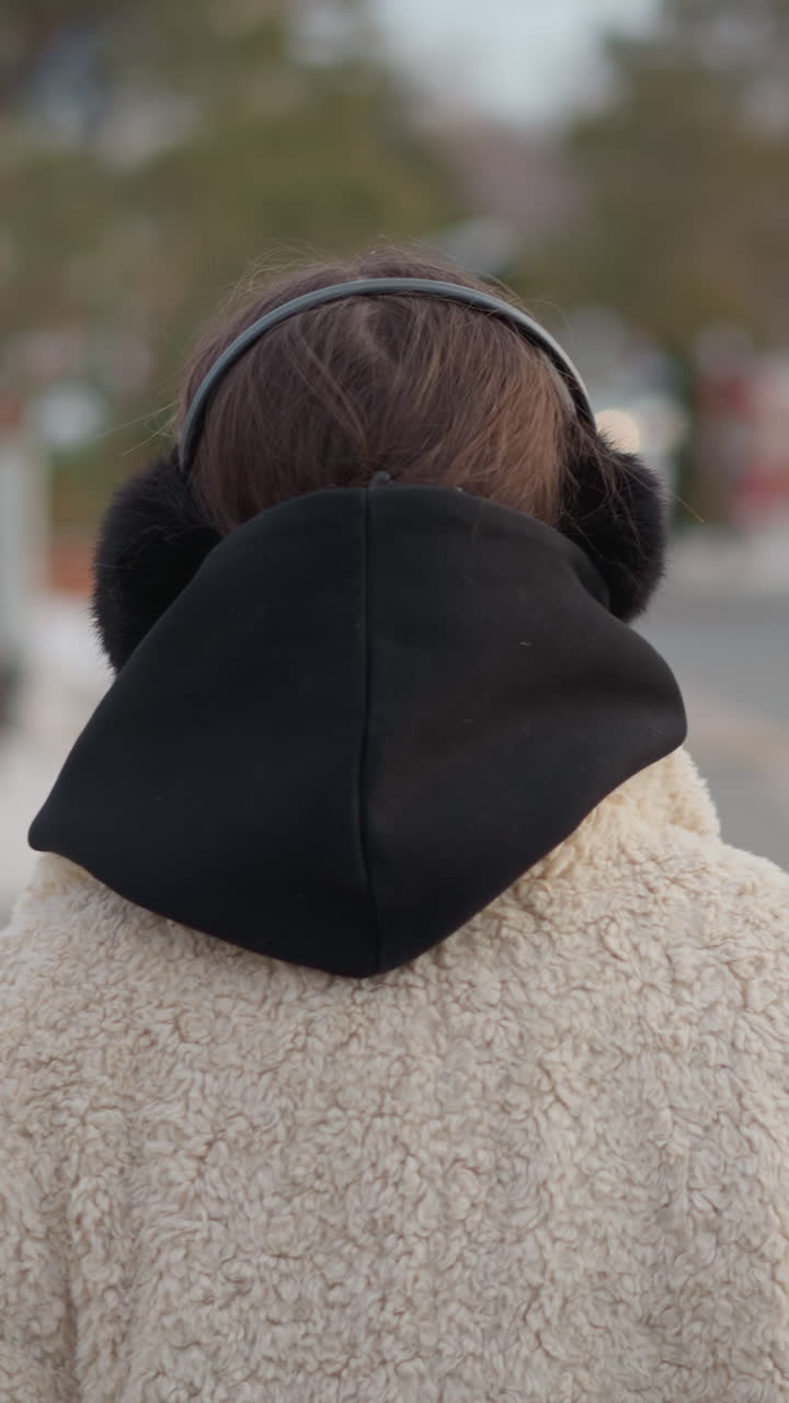 Persona con orejeras y abrigo de borreguito caminando por una calle urbana fría, con la capucha puesta, fondo suave con efecto bokeh, paleta de colores apagados, retrato trasero espontáneo con un ligero giro de cabeza, ropa de abrigo con textura que transmite
