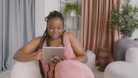 woman with braids lying on a big sofa while smiling and using a tablet