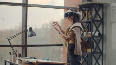 Moderne-Architektin-Im-Büro-Mit-Großen-Fenstern-Steht-In-Einem-Virtual-Reality-Helm-Und-Nutzt-Gesten,-Um-Das-Projekt-Zu-Verwalten,-Ohne-Das-Büro-Zu-Verlassen.-Baukontrolle.-Designprojekt-Des-Gebäudes-Und-Des-Innenraums