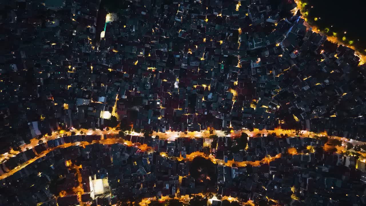 Top-down aerial of Hanoi, Vietnam, showing a dense urban layout illuminated by winding streets glowing with orange and white lights. The glowing roads carve through a compact grid of buildings