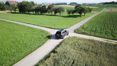 a volvo xc 90 car drives slowly before an intersection between two small roads before continuing straight ahead, swiss countryside and fields, aerial shot, drone