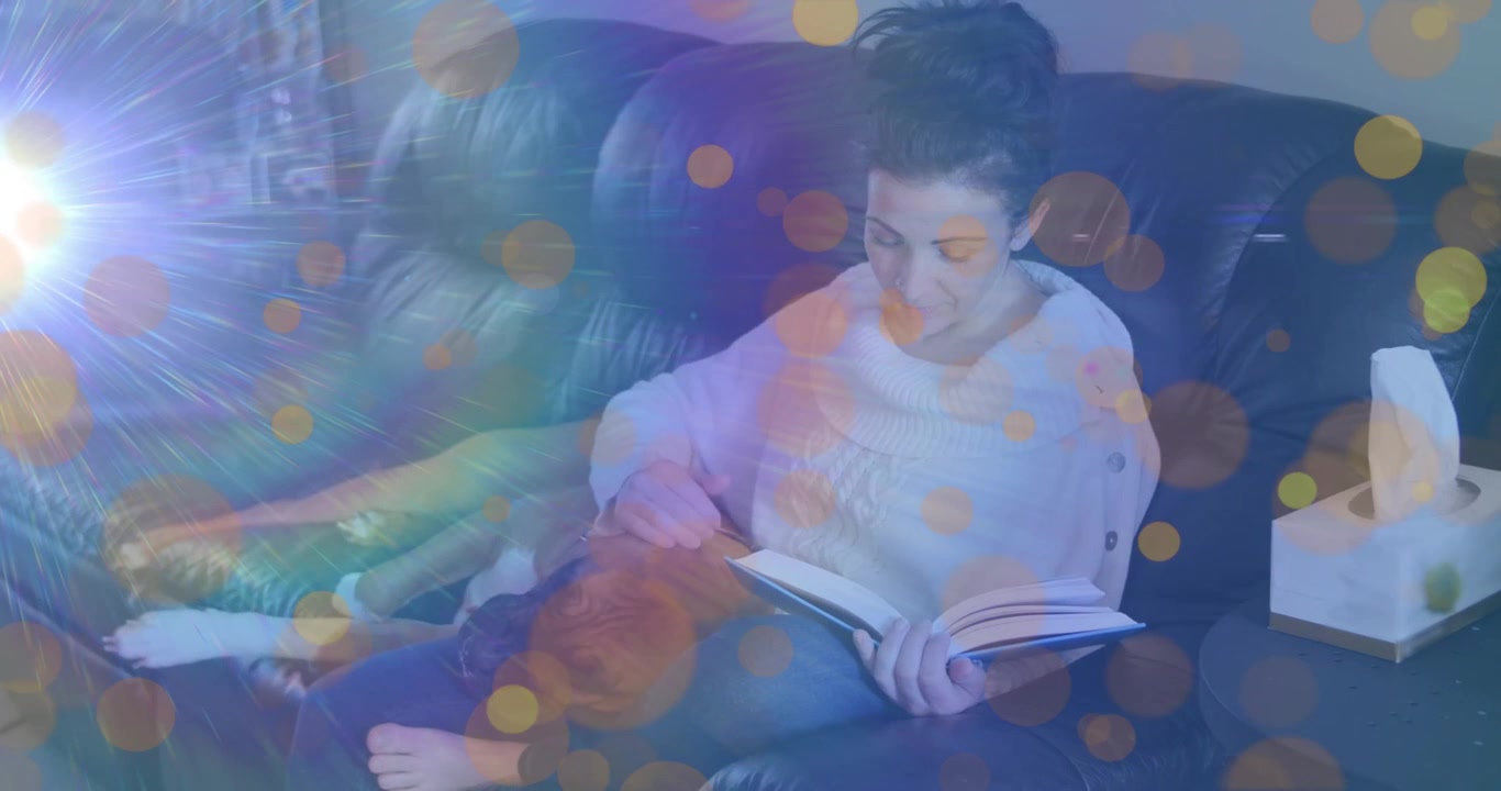Donna di mezza età che alterna la lettura di un libro sul divano e accarezza un cane che emerge da sotto una coperta, per studio.