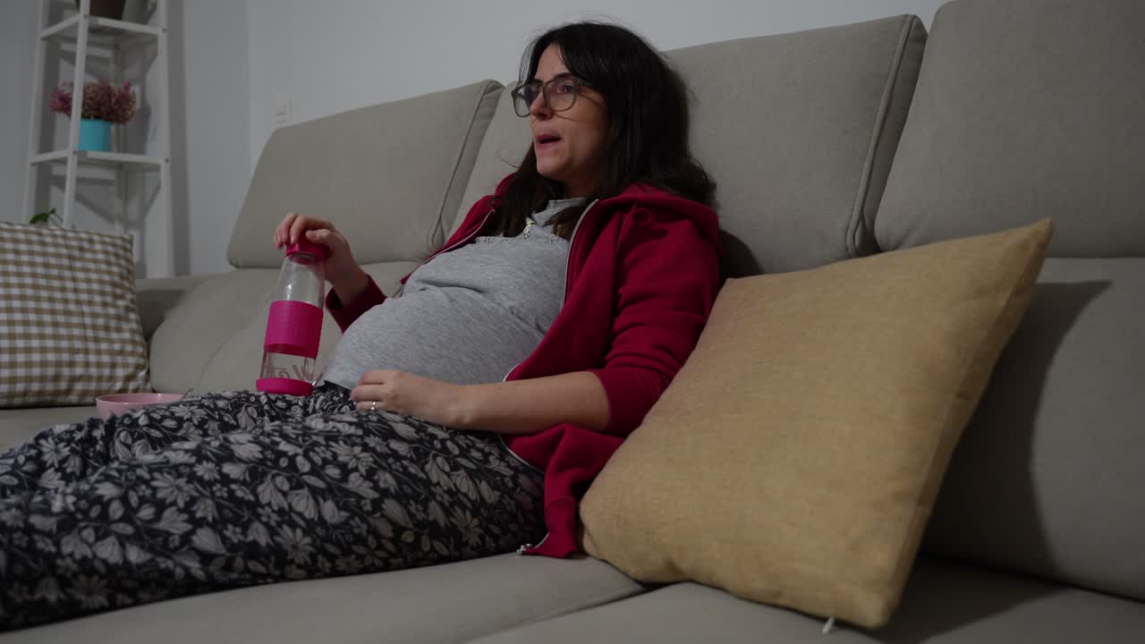 Pregnant woman watching TV and drinking water at night, relaxed on home sofa