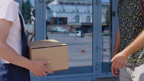 a delivery worker in a white t-shirt and blue coveralls hands the box to another male recipient near the glass modern front door of the high-rise. delivery of goods to the office