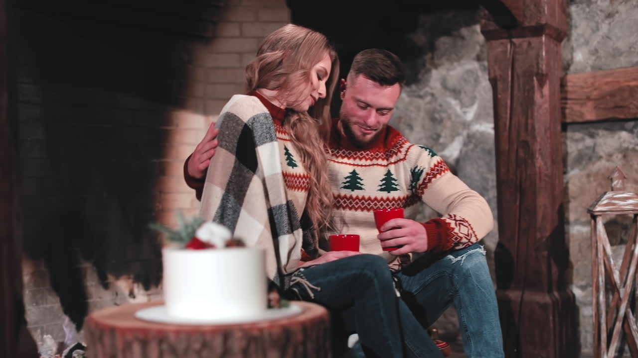Couple in love drinking tea. Young man and beautiful girl in knitted Christmas sweaters sitting near the fireplace. Man kisses his girlfriend. Romantic evening