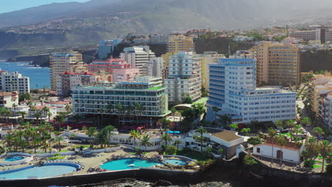luxurious hotels and touristic resorts with swimming pools and palms, in puerto de la cruz, canary islands, spain, and its harbor on the coast of the atlantic ocean, aerial rotating shot 4k