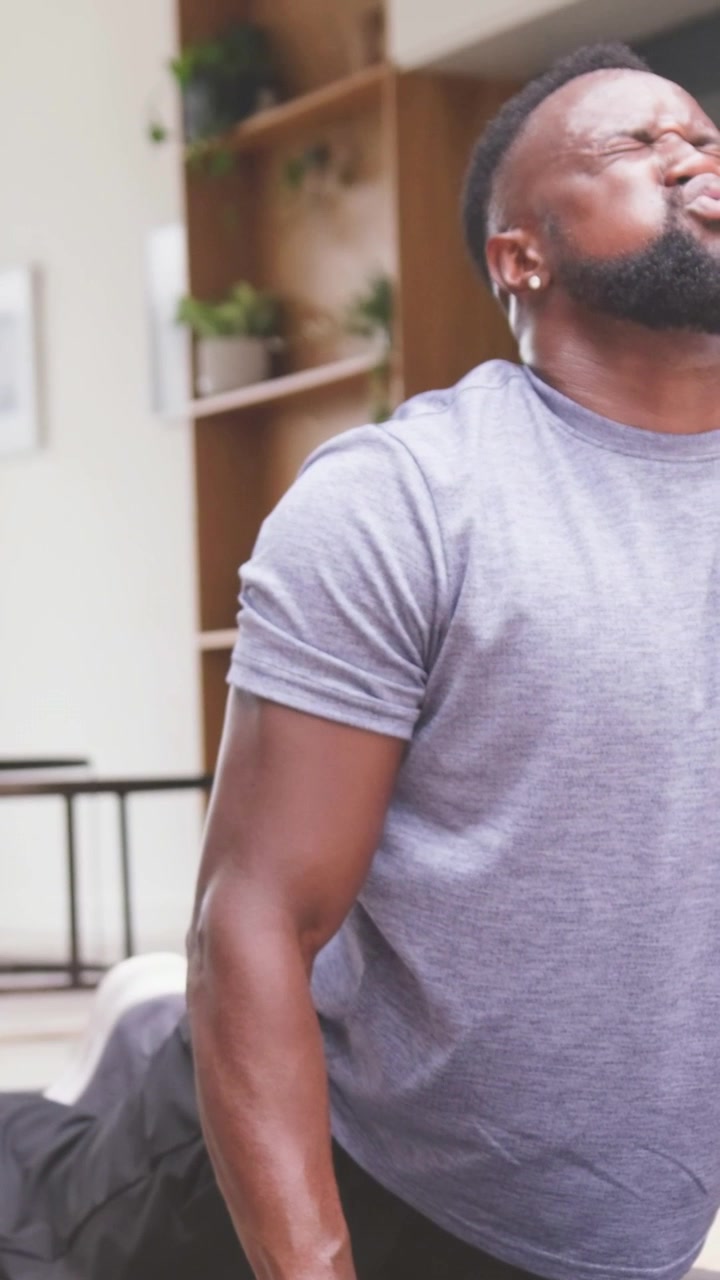 Vertical video: Exhaling strongly, man pressing up on yoga mat at home for back stretch in grey tee