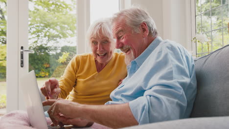 retired senior couple sitting on sofa at home using laptop online shopping or booking holiday