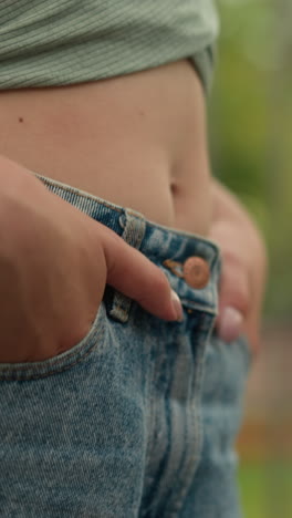 close-up of young woman walking with hands in pockets, exposing midriff, wearing green cropped top and high-waisted jeans, beige handbag on arm, with blur background
