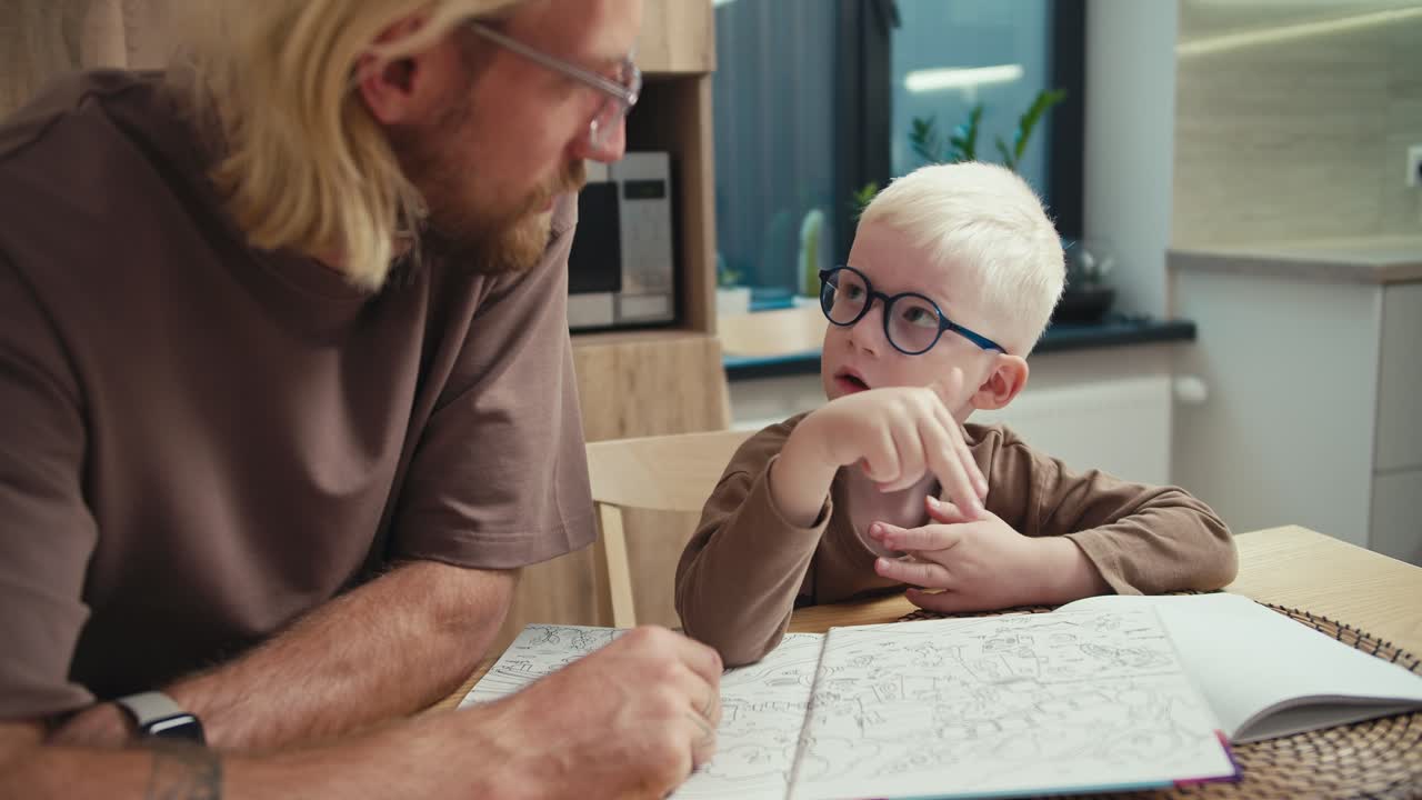 An interested albino boy with blue glasses and white hair communicates with his father, a blond man with a beard and glasses, about a certain topic at school and the boy wants to learn new things in the kitchen in a modern apartment with his father