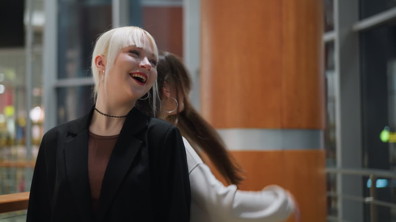Side view of girl standing in stylish outfit while sibling behind flaunts long hair near reflective glass indoors creating modern urban vibe with natural expressions youthful energy