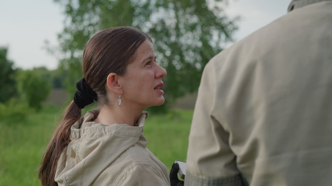 rear view of woman in field facing colleague engaged in conversation with gaze, hair pulled into ponytail, wearing jumpsuit and gloves, lush green forest backdrop