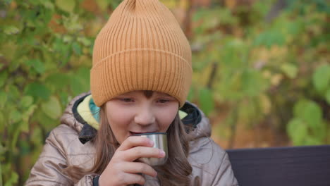close up of little girl in orange knit hat and bronze coat dipping finger into warm tea to test temperature before sipping, sitting on bench surrounded by vivid autumn leaves and gentle outdoor light