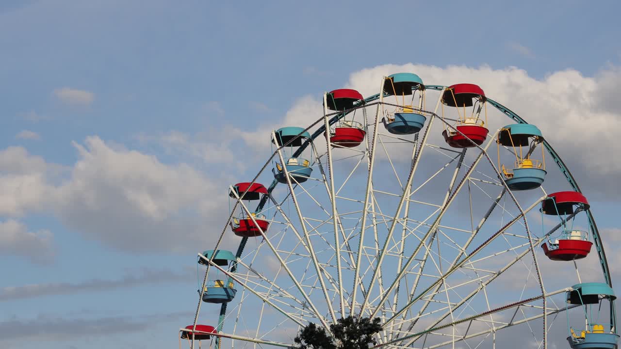Large ferris wheel with open cabins quickly turns against a blue sky