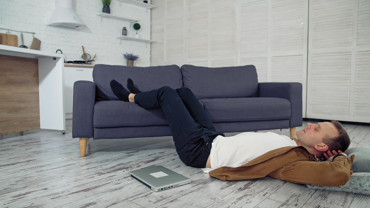 Tired freelance worker rests on floor. Young guy sleeping on the kitchen floor with his legs on sofa. Laptop lying on the floor. Remote job. Quarantine. Lockdown concept.