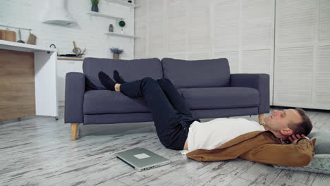 tired freelance worker rests on floor. young guy sleeping on the kitchen floor with his legs on sofa. laptop lying on the floor. remote job. quarantine. lockdown concept.