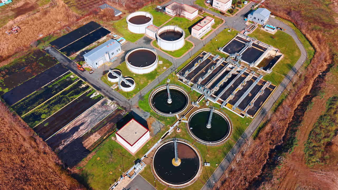 Overhead drone shot of a modern wastewater treatment plant. Clean layout of circular and rectangular water purification tanks with asphalt roads and green spaces