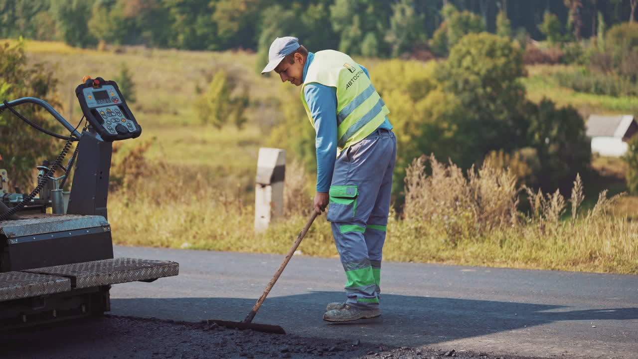 Workers make road asphalting on the natural background of trees. Male workers in protective uniform are working on one side of the road. New asphalt on the highway.