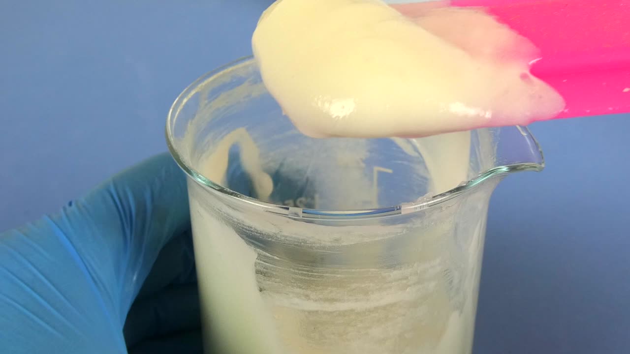 Macro shot of a cosmetic emulsion being prepared in a laboratory. The video captures the operator's gloved hands mixing ingredients in a beaker,