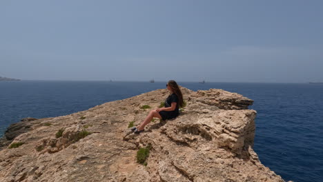 Woman-sits-down-leaning-up-against-rocky-ridgeline-looking-over-blue-ocean