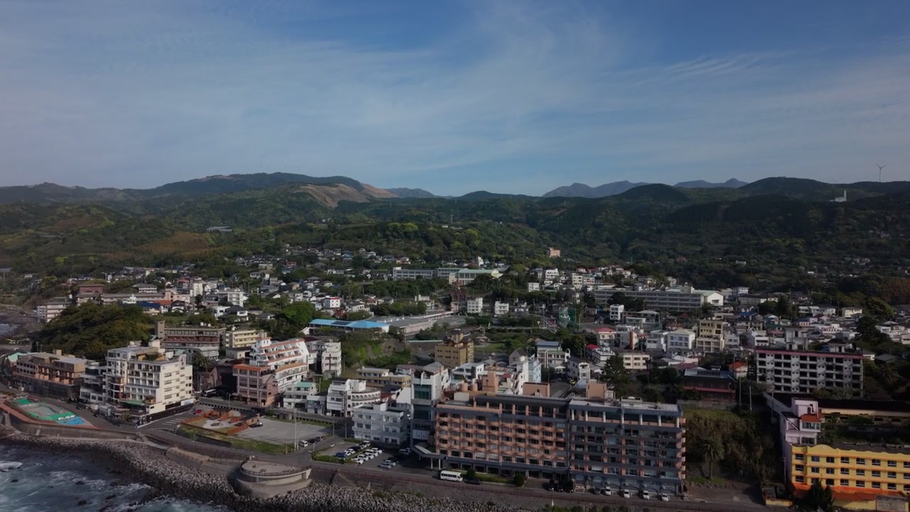 Multistory hotel buildings lining a rocky stretch of coastline below wooded hills, Higashiizu, Izu, Japan, under hazy afternoon light, aerial drone shot