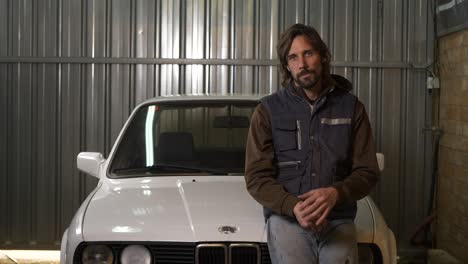 Man-in-mechanic-vest-sits-on-white-car-hood,-tosses-keys,-and-winks,-Slow-Motion