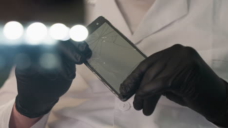 a close-up view of a technician in a white lab coat and black gloves examining a cracked smartphone screen